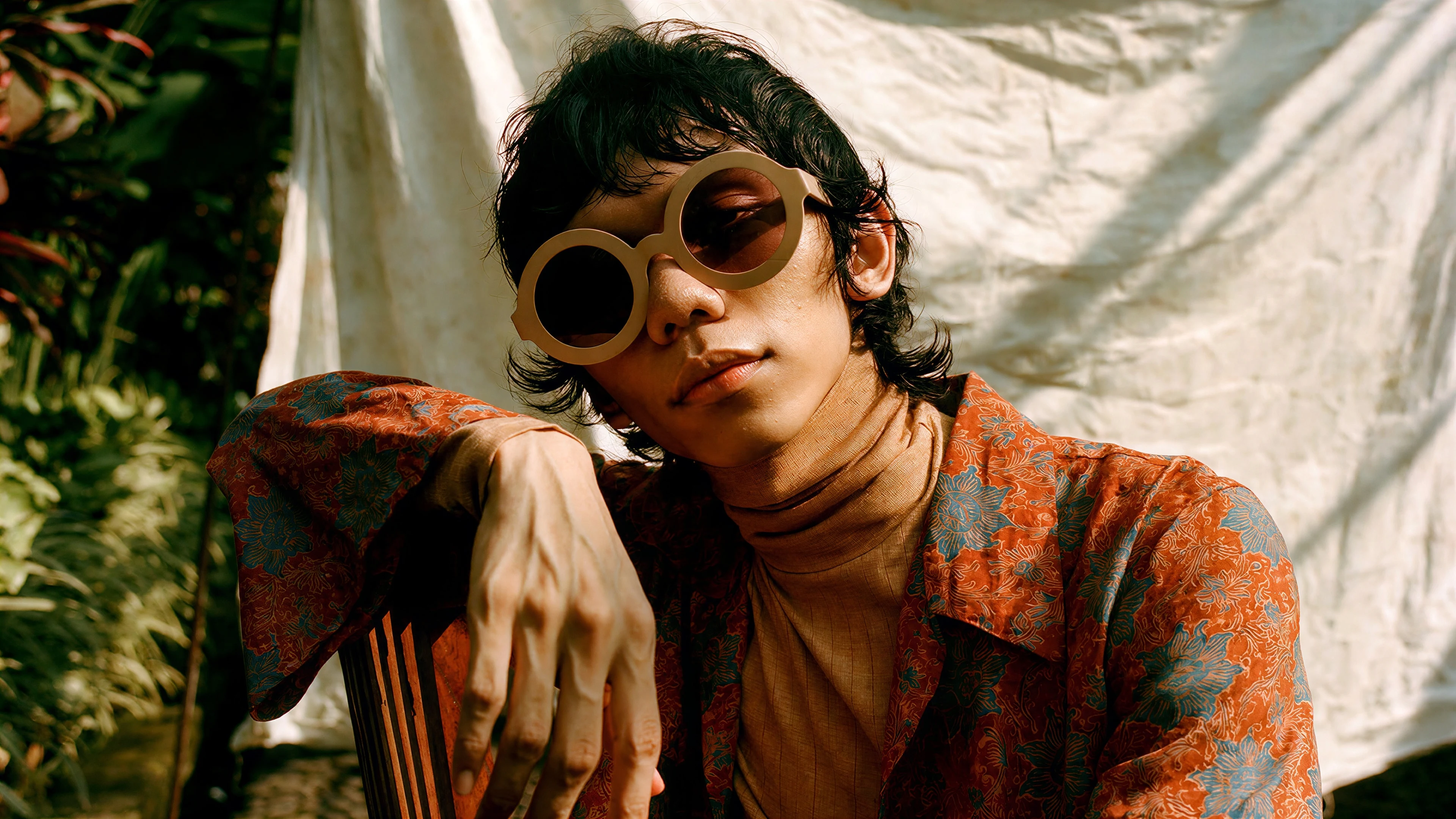 A man draped over a chair back in cream round sunglasses